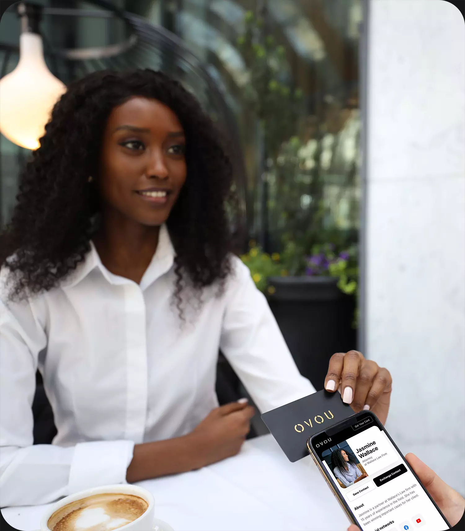 Black woman tapping her OVOU digital business card on prospects phone. Her OVOU profile is shown on mobile phone of recipient. Setting is high end / premium restaurant with fireplace on the right and cafe latte on the table.